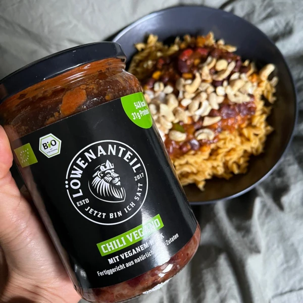 Hand hält Löwenanteil Bio Chili Vegan Glas neben Schüssel mit veganem Chili und Bohnen